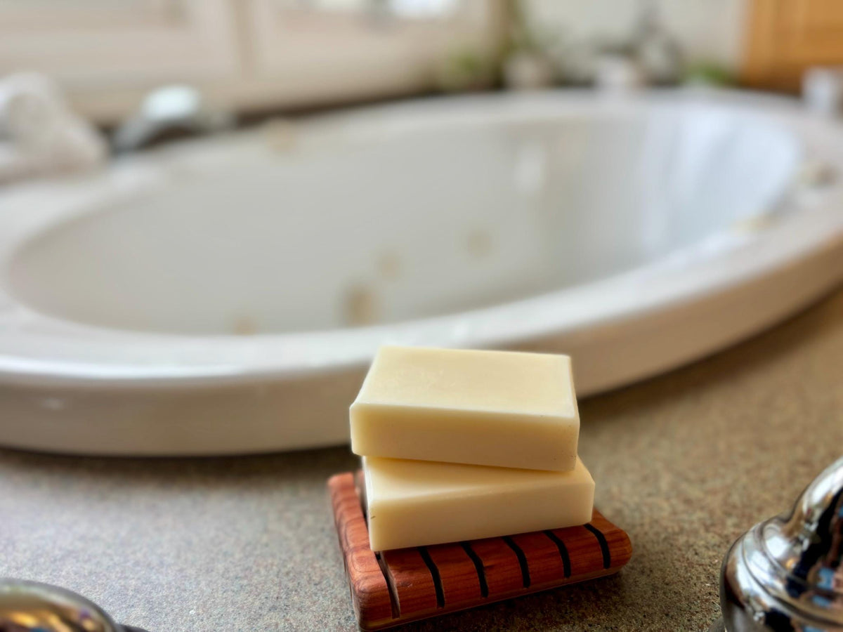 Two soap bars on a wooden soap dish with a blurred bathroom background