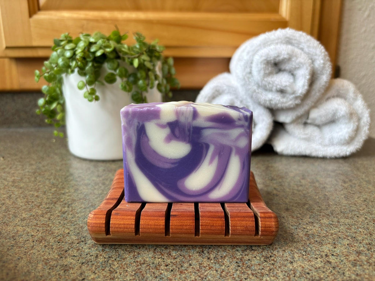 Purple and white soap bar on a wooden soap dish with towels and a plant in the background.