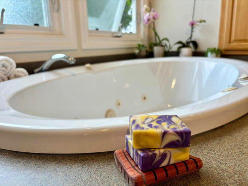 Stack of purple and yellow swirled soap bars on a wooden soap dish in front of a bathtub.