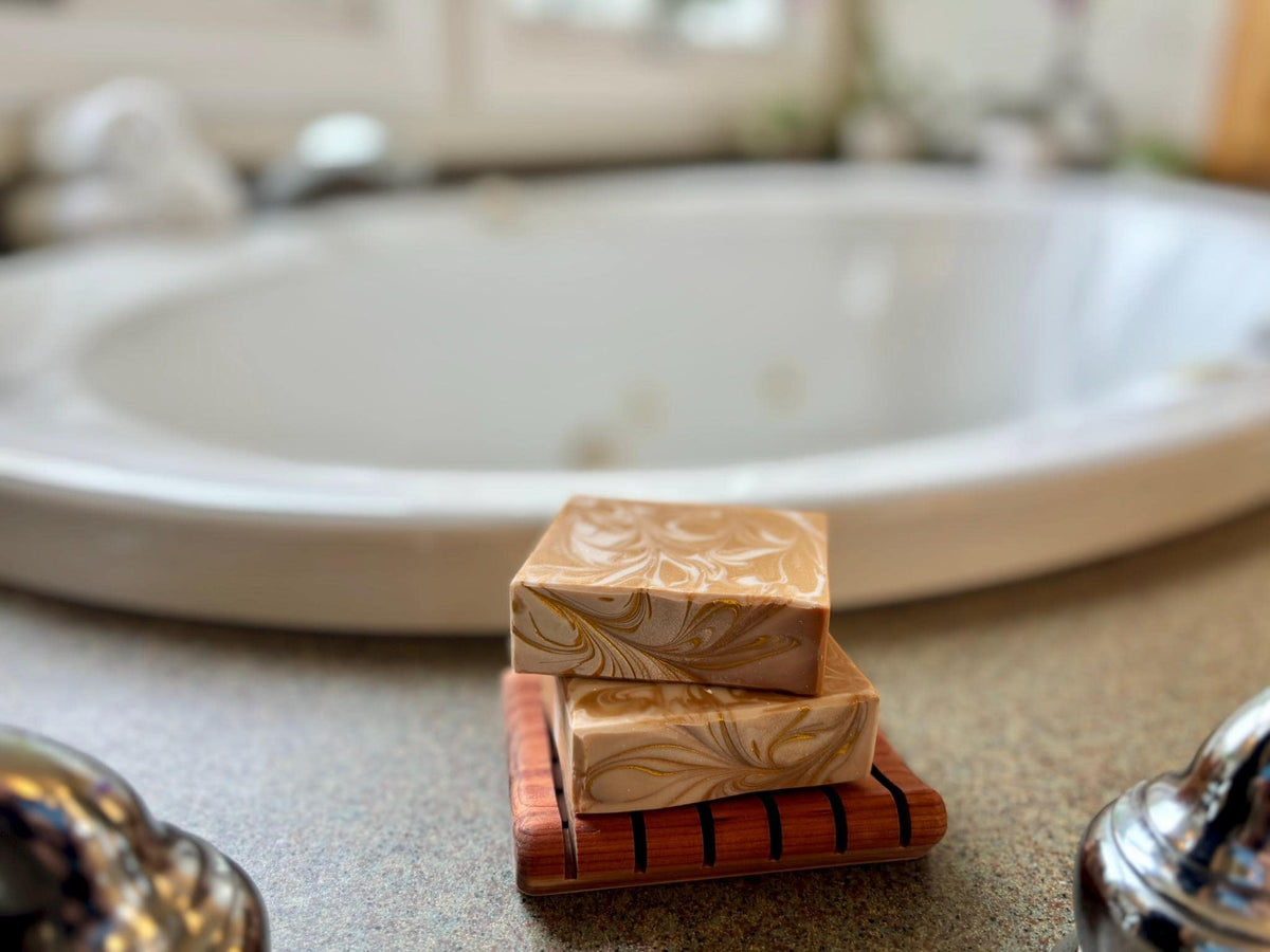Two soap bars on a wooden soap dish with a bathroom sink in the background