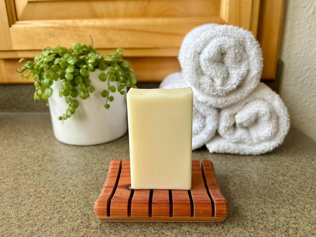 Bar of soap on a wooden soap dish with a towel and plant in the background