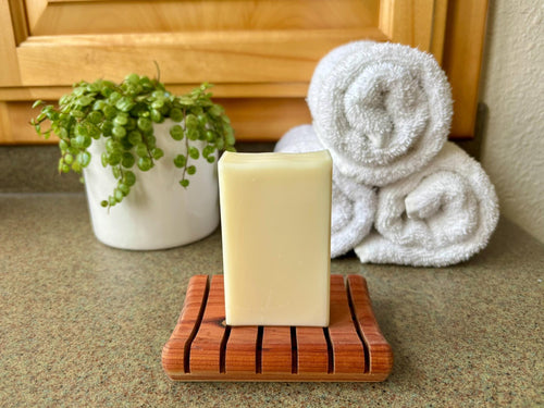 Bar of soap on a wooden soap dish with a towel and plant in the background