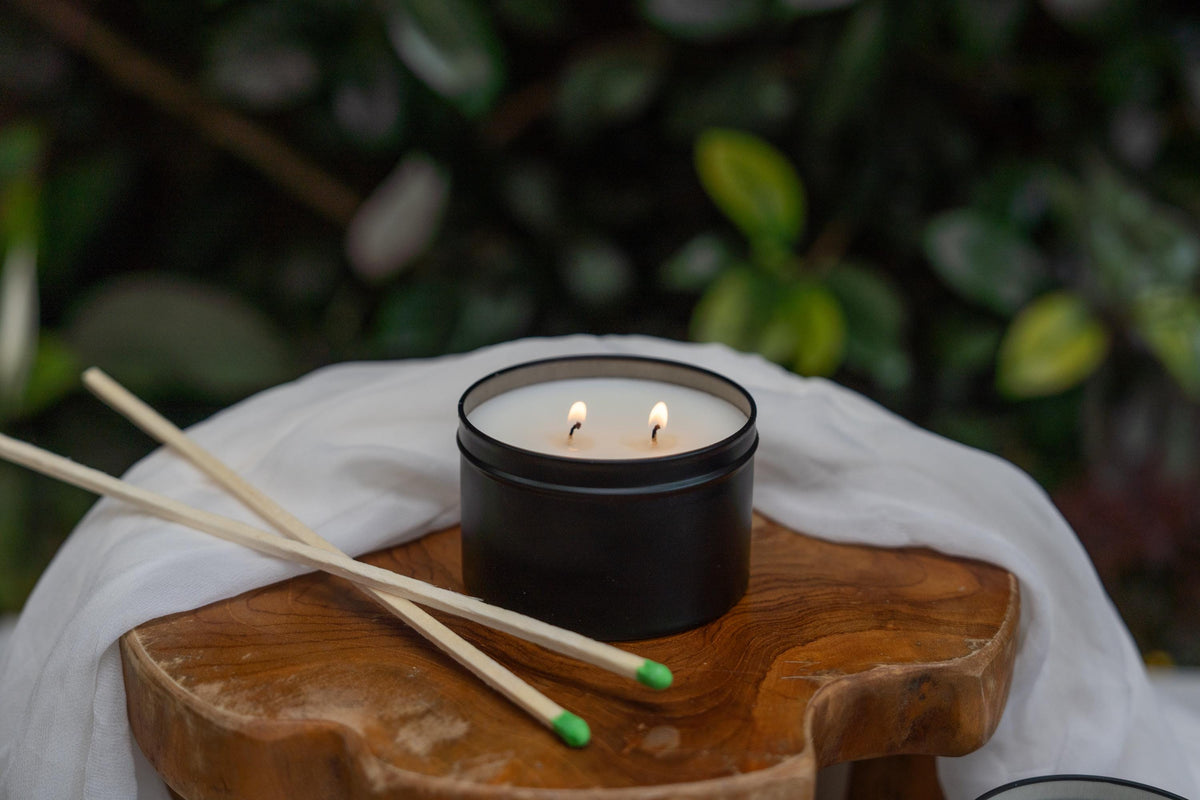 Black candle with two lit wicks on a wooden coaster, with two wooden match sticks beside it on a white cloth.