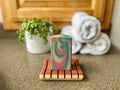 Colorful soap bar on a wooden dish with towels and a plant in the background