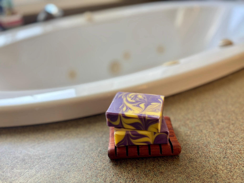 Two soap bars swirled with purple and on a wooden dish with a bathtub in the background.