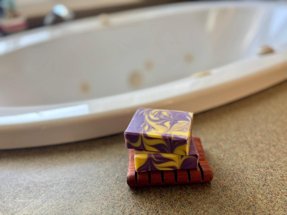Two soap bars swirled with purple and on a wooden dish with a bathtub in the background.