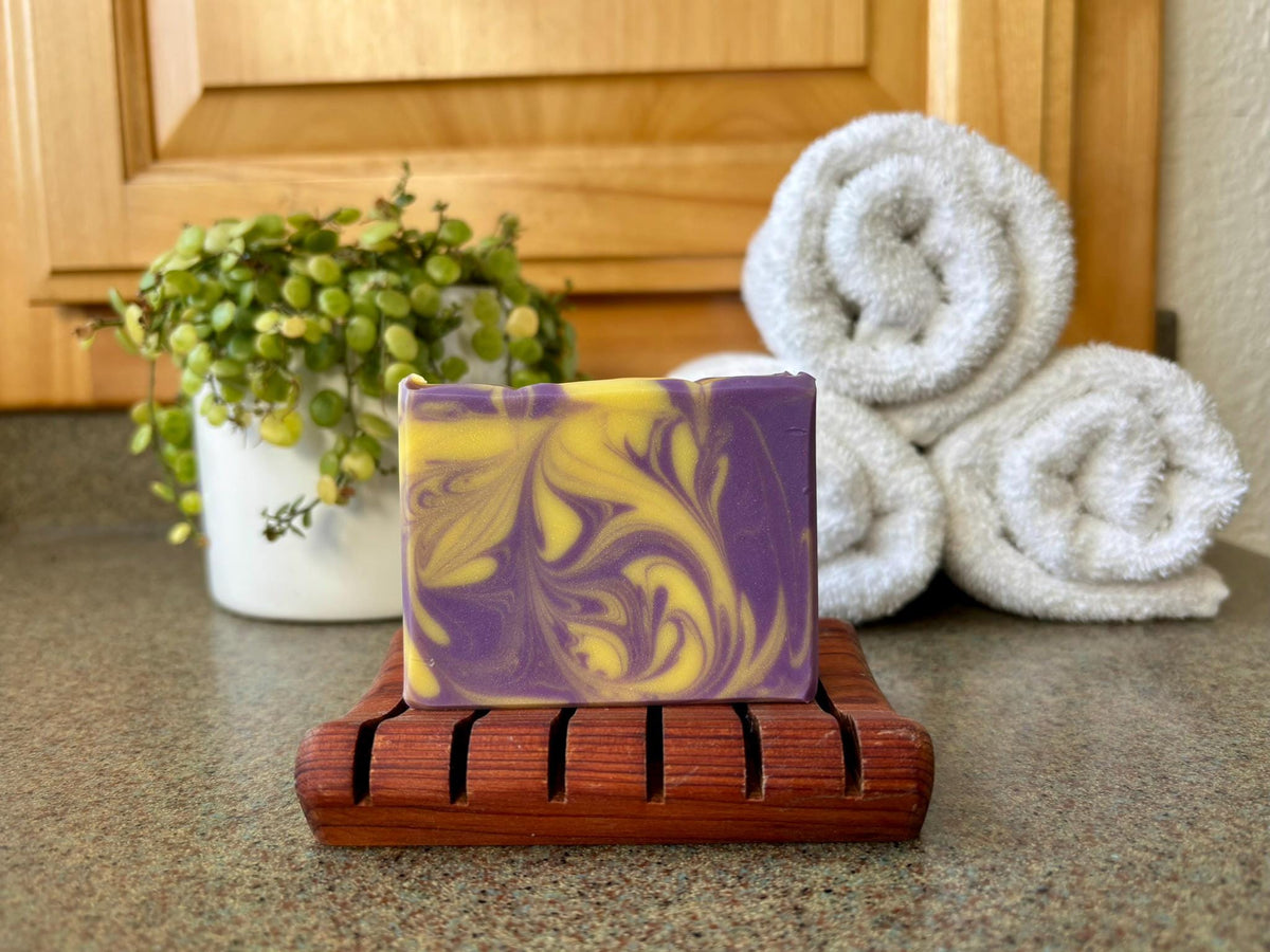 Purple and yellow soap bar on a wooden dish with white towels and a plant in the background.