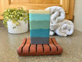 Bar of soap with gradient colors on a wooden soap dish, with a towel and plant in the background.